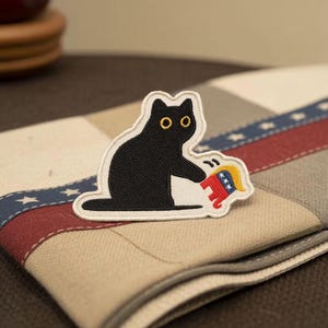 May include: Embroidered patch featuring a black cat with yellow eyes, holding a red, white, and blue emblem. The patch is on a beige and red striped fabric with blue star accents. The cat is the primary focus.