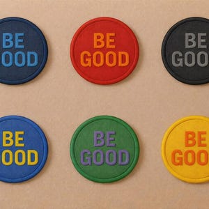 May include: Six circular patches in various colours, each bearing the words "BE GOOD". The patches are blue, red, black, green, and yellow. The text colour contrasts with the patch colour, creating a visually appealing design.