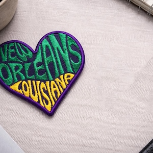 以下が含まれることがあります： 紫色の背景に、緑と黄色で「New Orleans Louisiana」と刺繍されたハート型のパッチ。 パッチは明るい色の表面にあり、背景にはノートと植物があります。
