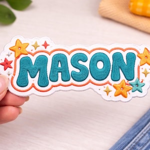 May include: Embroidered patch with the name "MASON" in teal bubble letters, outlined in orange, and surrounded by orange, teal, and red stars. The patch has a white border and is held by a hand.
