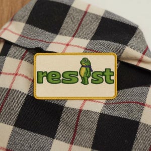May include: Embroidered patch with the word "resist" in green, featuring a cartoon frog wearing a purple scarf. The patch has a cream background and a gold border. The patch is placed on a black, white, and red plaid shirt.