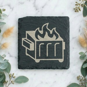 Op de afbeelding: Een vierkante zwarte leistenen onderzetter met een ruwe, onregelmatige rand. Het midden bevat een lichtgekleurde, gegraveerde afbeelding van een brandende afvalcontainer, weergegeven met eenvoudige lijnen en een vlammenicoon.