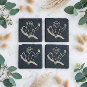 May include: A set of four square black slate coasters, each featuring an identical etched illustration of a monkey sitting on a tree branch. The coasters have a rough, natural stone texture and a dark grey finish with light-coloured monkey designs.