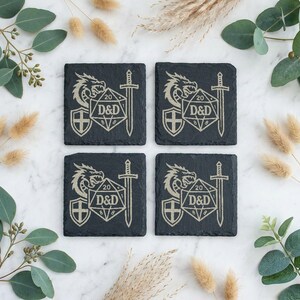 May include: Four square black slate coasters featuring an engraved design of a dragon, a shield, a sword, and a twenty-sided die with the text D&D. The coasters have a rough, natural edge texture and are arranged in a two-by-two grid pattern.