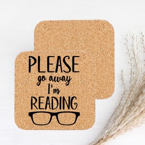 Puede incluir: Dos posavasos cuadrados de corcho. Uno tiene el texto negro "PLEASE go away I'm READING", con un gráfico de gafas negras. El otro posavasos es liso. Los posavasos están sobre una superficie blanca.