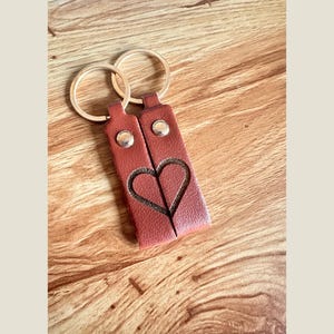 Puede incluir: Dos llaveros de cuero marrón rojizo a juego, cada uno con un recorte de medio corazón que forma un corazón completo cuando se juntan. Cuentan con remaches plateados y aros dorados.