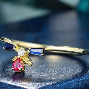 May include: A gold ring featuring a V-shaped band with a central design resembling a bee. The design includes two dark blue baguette-cut stones for wings, a small clear round stone for the head, and a vibrant red pear-cut stone for the body.