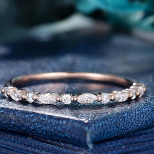 Könnte beinhalten: Ein roségoldener Bandring mit abwechselnd runden und marquiseförmigen, klaren Edelsteinen. Der Ring liegt auf einer dunkelblauen, strukturierten Oberfläche mit verschwommenem Hintergrund.