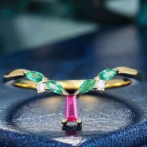 May include: A gold V-shaped ring featuring a central vertical baguette-cut pink gemstone, flanked by four marquise-cut green gemstones and two small round clear stones arranged in a symmetrical pattern along the band's curved top edge.