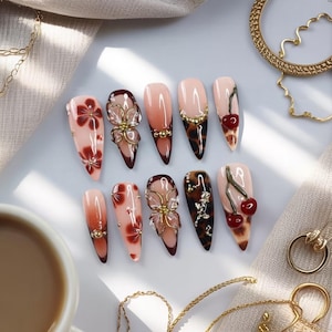May include: A collection of ten long, almond-shaped artificial nails. The nails feature various designs, including floral patterns, gold embellishments, and cherry accents. The color palette includes shades of pink, brown, and gold, creating a glamorous aesthetic.
