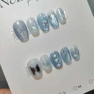 May include: Two sets of light blue press-on nails with various designs, including stars, butterflies, and abstract patterns. The nails are displayed on a white card with the text "NAIL BOX for you". Sizes XS, S, M, and O are indicated.