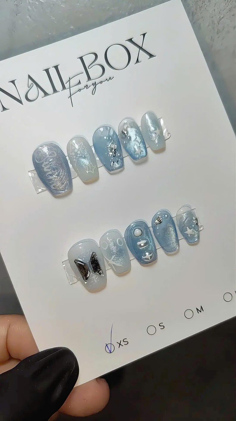 May include: Two sets of light blue press-on nails with various designs, including stars, butterflies, and abstract patterns. The nails are displayed on a white card with the text "NAIL BOX for you". Sizes XS, S, M, and O are indicated.