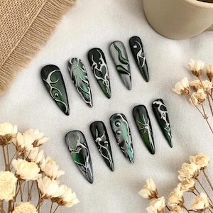 Könnte beinhalten: Ein Set aus zehn langen, mandelförmigen Kunstnägeln. Jeder Nagel ist mit einer dunkelgrünen Basis bemalt und weist komplizierte Designs in Weiß und Silber mit Akzenten in Grün und Grau auf. Die Designs variieren, einschließlich Wirbeln, Linien und abstrakten Mustern.