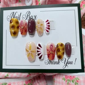 Può includere: Un set di dieci unghie finte esposte su un cartoncino bianco con le scritte Nail Box e Thank You. Le unghie presentano vari design, tra cui macchie di leopardo gialle e marroni, viola scuro traslucido, bianco con motivi rossi e fiori.