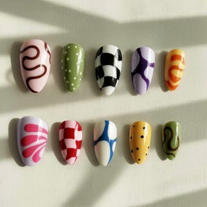 May include: A collection of ten artificial nails with various designs. The nails feature patterns such as polka dots, checkerboard, swirls, and abstract shapes in colors like pink, green, black, white, purple, orange, red, and yellow.