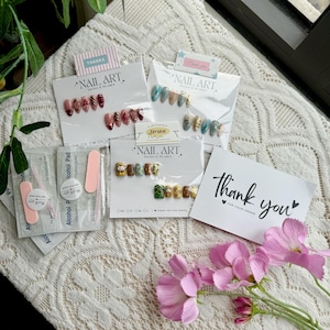May include: An assortment of nail art sets, each packaged on a white card with the words "NAIL ART" and "Thank you". The sets feature various designs, including floral, abstract, and character-themed nail art. Pink nail files and alcohol pads are also included.