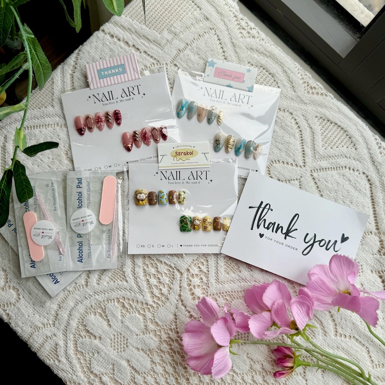 May include: An assortment of nail art sets, each packaged on a white card with the words "NAIL ART" and "Thank you". The sets feature various designs, including floral, abstract, and character-themed nail art. Pink nail files and alcohol pads are also included.