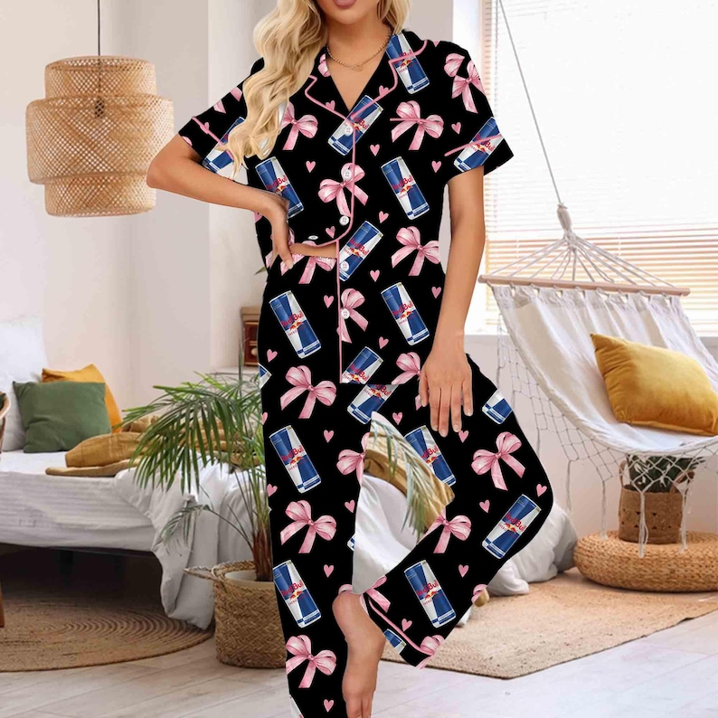 Puede incluir: Conjunto de pijama negro con un estampado de latas de Red Bull, lazos rosas y corazones. La parte superior de manga corta y los pantalones tienen ribetes rosas. El conjunto de pijama est&aacute; hecho de un material suave y c&oacute;modo.