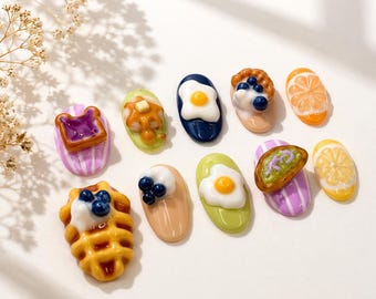 Könnte beinhalten: Eine Sammlung von Miniatur-Nageldesigns zum Frühstücksthema. Die Designs umfassen eine Waffel mit Blaubeeren, Toast mit Marmelade, Spiegeleier und Obstscheiben. Die Nagelkunststücke sind in verschiedenen Farben und Formen gehalten und haben ein glänzendes Finish.