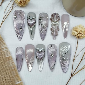 May include: A set of ten artificial nails in diverse designs. The nails display silver, grey, and pink hues, with detailed decorations. Some nails have heart-shaped designs, while others feature abstract patterns and glitter. Measurements are not applicable.