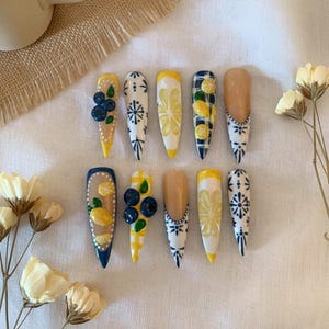 May include: A set of ten long, almond-shaped artificial nails. The nails feature various designs, including lemons, blueberries, and floral patterns in yellow, blue, and white. Some nails have a nude base with detailed artwork, while others are solid colors.