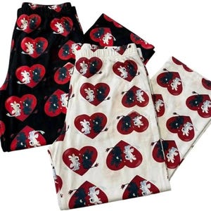 Puede incluir: Tres pares de pantalones de pijama con un patrón repetido de corazones rojos y dragones de dibujos animados. Un par es negro, otro es blanquecino y el tercero es negro. Los dragones son blancos y negros.