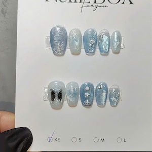 May include: A set of light blue press-on nails with various designs, including glitter, stars, and a bow. The text "NAIL BOX for you" is visible on the card. Sizes XS, S, M, and L are indicated.