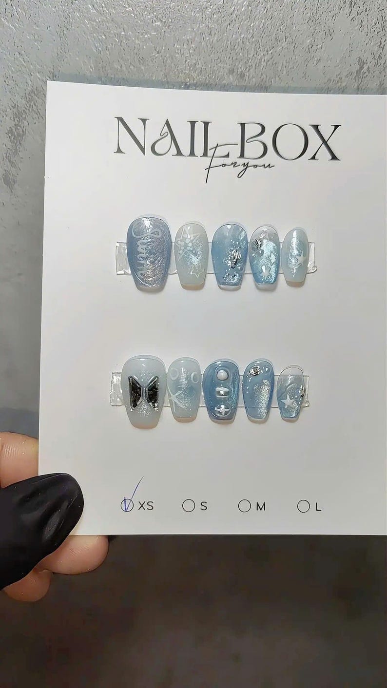 May include: A set of light blue press-on nails with various designs, including glitter, stars, and a bow. The text "NAIL BOX for you" is visible on the card. Sizes XS, S, M, and L are indicated.