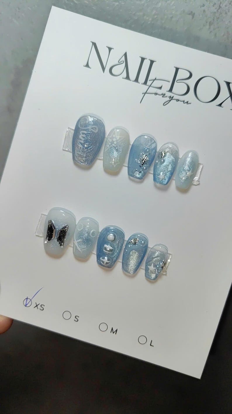 May include: Two sets of light blue press-on nails with various designs, including glitter, stars, and a butterfly. The top of the card reads "NAIL BOX for you". Sizes XS, S, M, and L are indicated.