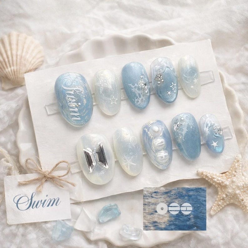 May include: A set of ten artificial nails in various shades of blue and white, with decorative elements. Some nails feature the word "Swim", stars, and silver embellishments. The nails are arranged on a white surface with a seashell and starfish.