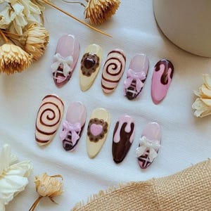 Pode incluir: Um conjunto de dez unhas postiças em forma de amêndoa com vários designs. As unhas apresentam uma mistura de cores rosa, castanho e creme, adornadas com laços, corações e padrões de redemoinho. Algumas unhas têm um design de gotejamento de chocolate.