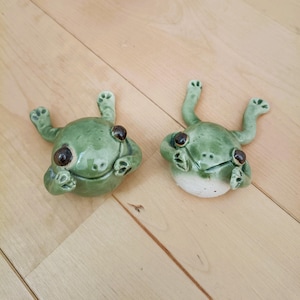 Puede incluir: Dos figuras de rana de cerámica en un esmalte verde claro con ojos marrones. Las ranas están tumbadas boca arriba con los brazos y las piernas extendidos. Las figuras tienen un aspecto artesanal y caprichoso.