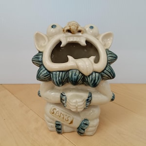 Könnte beinhalten: Cremefarbene Keramikfigur einer Fabelwesen mit offenem Mund. Die Figur hat blaue Akzente an Mähne und Füßen, und der Text "SAM'S" ist auf der Vorderseite zu sehen.