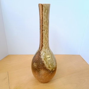以下が含まれることがあります： 細長い首と球根状の底を持つ背の高い陶器製の花瓶。表面は茶色、黄褐色、クリーム色の土のような色合いの斑点のある釉薬で仕上げられています。花瓶の側面に日本語のテキストが書かれた小さな明るい色のステッカーが貼られています。
