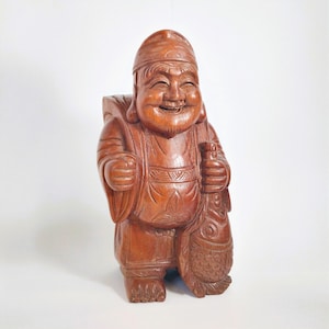 May include: A carved wooden statue of a smiling man wearing a hat and carrying a bag and a fish. The statue is brown and has detailed carvings, with a joyful expression on the face. The figure is standing upright.