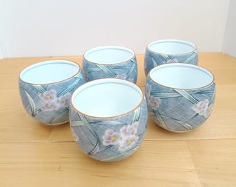 Könnte beinhalten: Ein Set aus fünf Teetassen aus Keramik mit einem blau-weißen Kreuzschraffurmuster, hellvioletten Blumenakzenten und grünen Blattmotiven. Jede Tasse hat eine glatte, abgerundete Form mit einem dünnen braunen Rand und weißem Inneren.