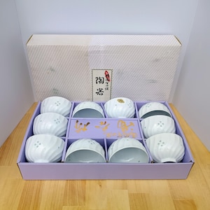 May include: A set of ten white ceramic bowls with a spiral ribbed texture and small cut-out patterns near the rim, arranged in a light purple cardboard tray inside a white box featuring Japanese text and a decorative label on the lid.