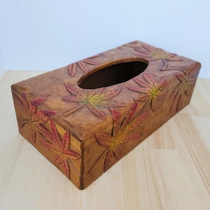 May include: A rectangular wooden tissue box with carved maple leaf designs. The leaves are painted with red, yellow, and green hues. The box has a dark brown oval opening on top for tissue dispensing.
