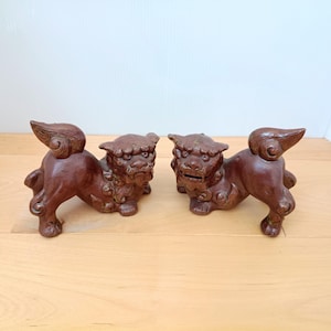 Puede incluir: Un par de figuras de leones guardianes de metal fundido de color marrón oscuro. Cada criatura está representada en una postura agachada con melenas estilizadas, colas rizadas y rasgos faciales detallados. Un león tiene la boca cerrada y el otro abierta.