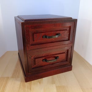 May include: A small, two-drawer wooden cabinet with a rich, reddish-brown finish. The cabinet features rectangular drawers with dark metal handles. The top of the cabinet is flat, and the wood grain is visible throughout.