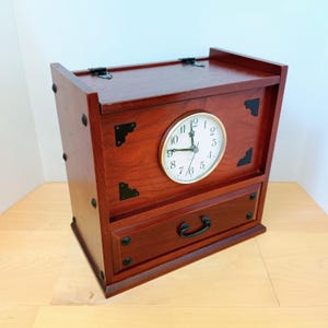 Puede incluir: Un cofre rectangular de madera con acabado marrón rojizo que presenta una esfera de reloj analógica circular con números y manecillas negras. El cofre incluye un cajón con un tirador de metal negro y detalles decorativos en las esquinas.