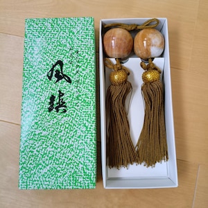 May include: A pair of polished, spherical, tan and white stone ornaments with long, dark gold-colored tassels, presented in a white box next to a green and white patterned lid featuring black Japanese calligraphy and text.