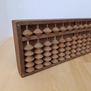 May include: A wooden abacus featuring a rectangular frame with vertical metal rods. Each rod holds multiple light brown, diamond-shaped beads. A horizontal divider separates the beads, with one bead above and four beads below on each rod.
