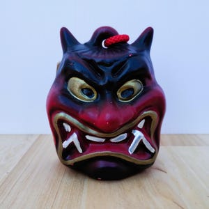 May include: A red, black, and gold Japanese demon mask with a fierce expression. The mask features horns, a wide mouth with sharp teeth, and gold-colored accents around the eyes and mouth. A red cord is attached at the top.