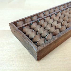 May include: A rectangular wooden abacus featuring a dark brown frame and multiple rows of smooth, rounded wooden beads mounted on metal rods. The beads are arranged in columns and rows, designed for manual calculation and counting tasks.