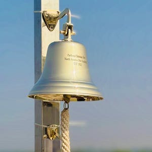 May include: Two shiny, silver-colored bells are mounted on a metal pole. The top bell has text that reads "Parfums Christian Dior North America Dior Academy EST 2025." A thick rope hangs from the top bell to the bottom bell.