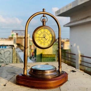 May include: A brass-toned metal desk clock suspended from an arched frame on a dark wooden base. The clock face is yellow with black Roman numerals and features the text Victoria London 1876 with an anchor graphic in the center of the dial.