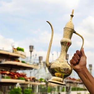 Puede incluir: Una jarra de metal dorado y ornamentada, con un pico largo y curvo y un asa. La jarra tiene grabados detallados y se sostiene contra un cielo nublado. El diseño sugiere una estética de Oriente Medio o antigua.