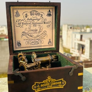 May include: A vintage nautical instrument set in a wooden box. The box is open, revealing a brass telescope and other instruments. The box has a label that reads "J. Scott London 1753". The box also has a label that reads "Mathematical And Nautical Instrument Maker".