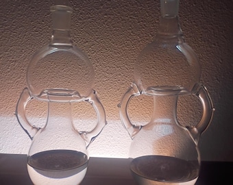 May include: Two clear glass vessels with double-chambered bodies and two side handles each. The vessels feature a rounded base and a smaller upper bulbous section, topped with a matching glass stopper. Both containers hold a small amount of liquid.